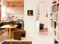 Un salón-cocina acogedor con estanterías llenas de objetos, un sofá cama y una mesa amarilla. Un pasillo arqueado conduce a otra estancia con una tabla de surf. Estas casas curiosas invitan a la vida.