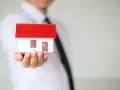 Un hombre con corbata negra sostiene una maqueta de casa blanca con techo rojo. ¿Listo para que puedas alquilar mi vivienda habitual?