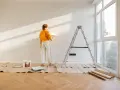 Mujer pintando pared con rodillo, lista para decorar piso antiguo de alquiler. Escalera, cubeta de pintura y protectores de suelo.