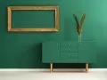 Un mueble verde esmeralda con patas doradas y un marco dorado vacío sobre pared verde. Los colores que pegan con el dorado son el verde intenso y el blanco.