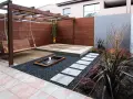 Un columpio de madera cuelga bajo un cenador. Senderos de piedra cruzan grava negra hacia un estanque. Minimalistas jardines modernos.