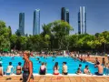 Gente disfrutando en una vibrante piscina comunitaria, con rascacielos modernos de fondo y árboles frondosos.
