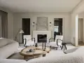 Sofá blanco texturizado, sillones blancos con brazos oscuros, mesa de centro de madera y ratán, chimenea y arte abstracto. Colores que combinan con el blanco crean un ambiente sereno.