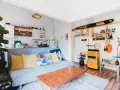 Sala de estar lista para alquilar a estudiantes. Sofá azul, mesa de centro de madera, estanterías con objetos decorativos y cocina pequeña.