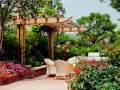 Jardín con pérgola de madera, mesa y sillas, flores rosas y rojas, y barbacoa.