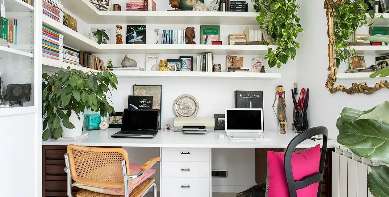 Un despacho en casa con estanterías llenas de libros y plantas. Dos portátiles, una impresora y decoración variada crean un espacio de trabajo funcional y acogedor.