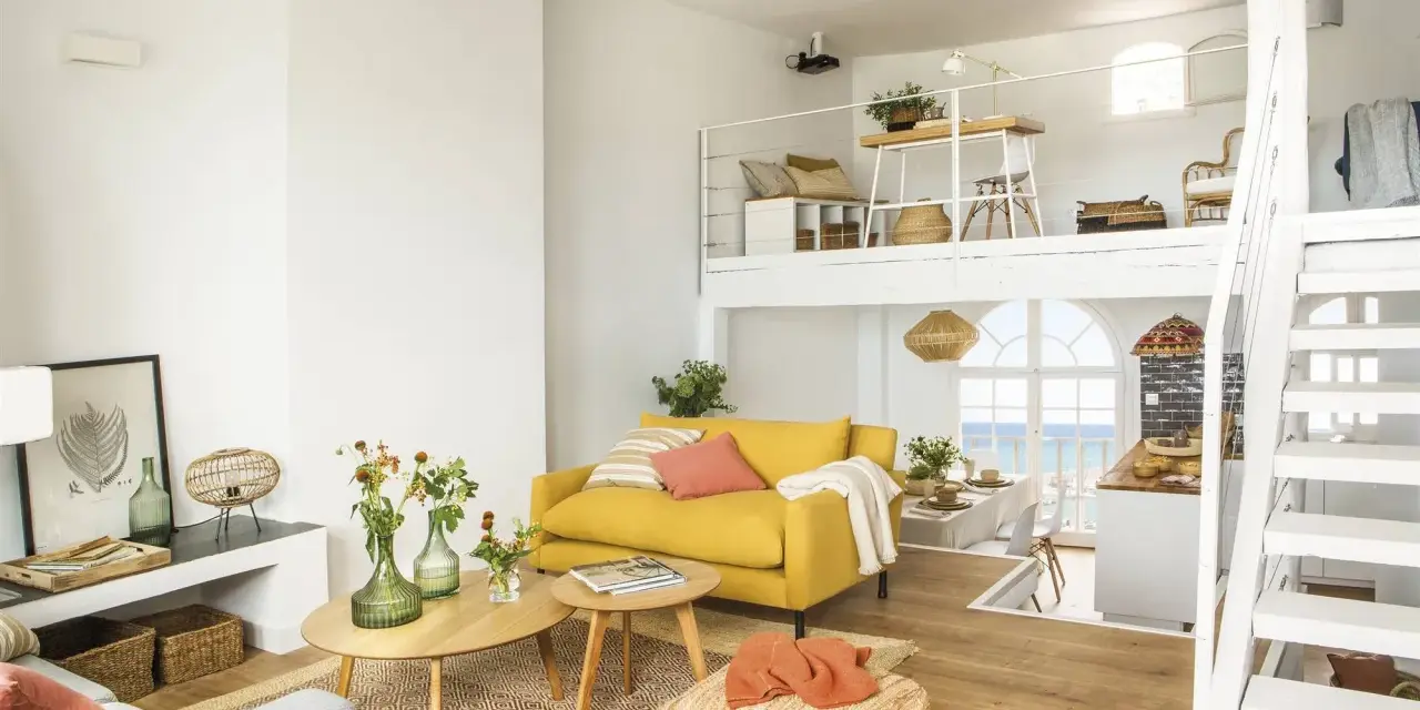 Un salón luminoso con sofá amarillo, mesas de madera y una escalera blanca que lleva a un altillo. Parece uno de esos **casas de cuento** con vistas al mar.