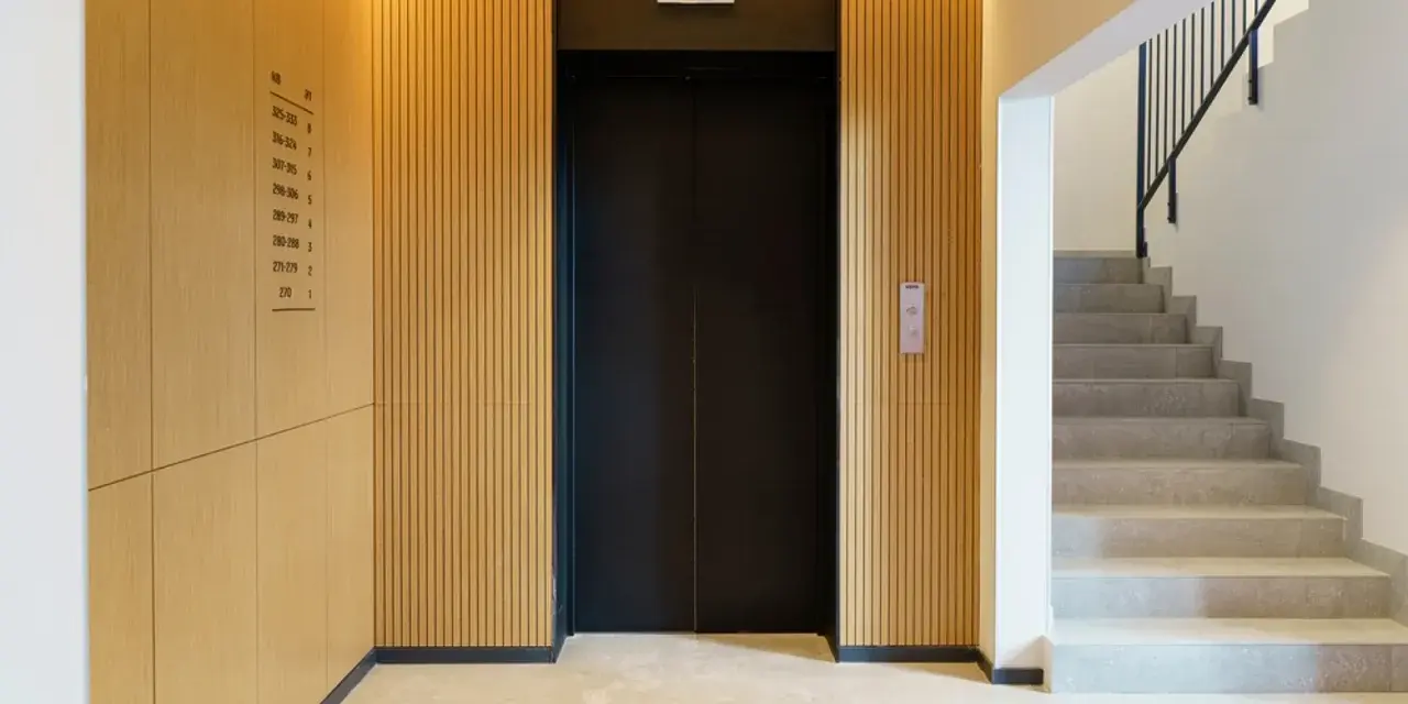Portal moderno con ascensor negro y escaleras de mármol. Ideas para como decorar un portal de vecinos con paneles de madera y luz cálida.