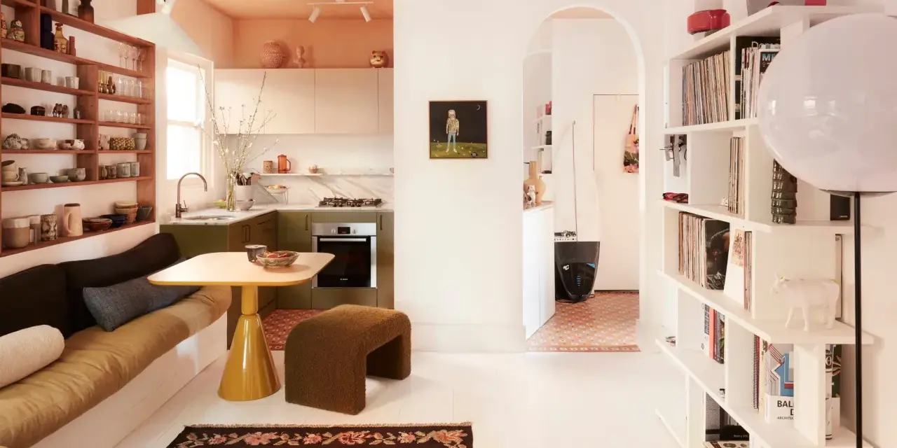 Un salón-cocina acogedor con estanterías llenas de objetos, un sofá cama y una mesa amarilla. Un pasillo arqueado conduce a otra estancia con una tabla de surf. Estas casas curiosas invitan a la vida.