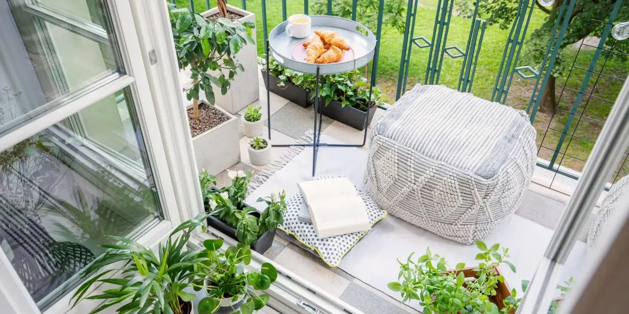 Acogedor rincón en unas terrazas cerradas pequeñas, con plantas, cojines, un libro y un café con cruasanes.