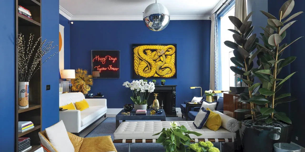 Salón moderno con paredes azul cobalto. Sofá blanco, sillón mostaza y cojines amarillos. Arte abstracto y plantas. Colores que combinan con el azul resaltan.