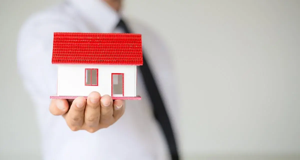 Un hombre con corbata negra sostiene una maqueta de casa blanca con techo rojo. ¿Listo para que puedas alquilar mi vivienda habitual?