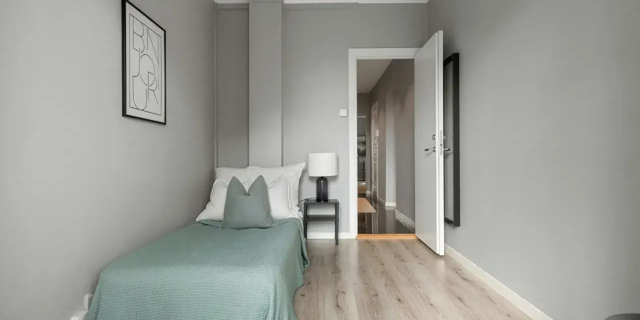 Habitación minimalista con cama individual, mesita de noche y lámpara. La puerta abierta revela un pasillo, sugiriendo que es legal alquilar habitaciones.