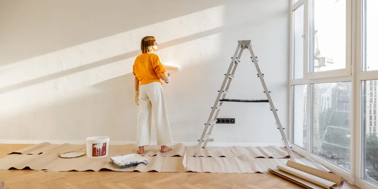 Mujer pintando pared con rodillo, lista para decorar piso antiguo de alquiler. Escalera, cubeta de pintura y protectores de suelo.