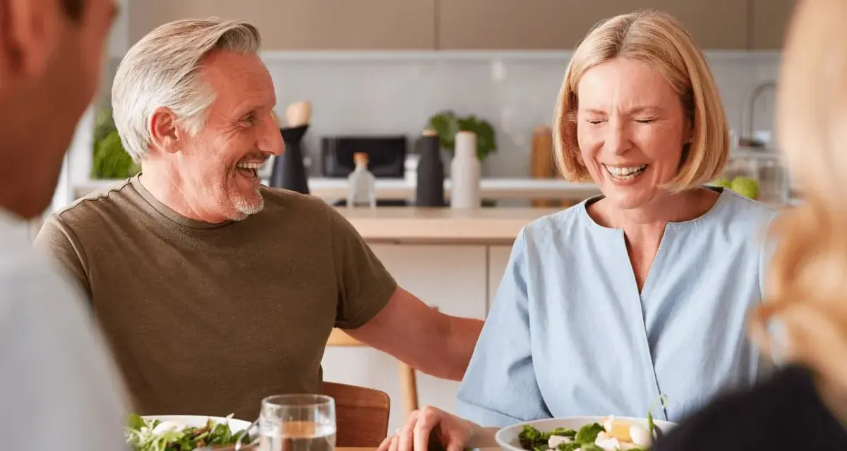 Padre e hijo ríen mientras planean comprar vivienda a medias. Comida familiar en la cocina.