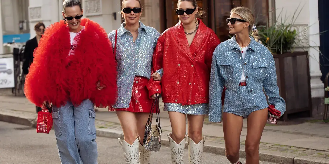 Cuatro mujeres pasean con atuendos llamativos. Una lleva un abrigo rojo voluminoso, otra un conjunto de lentejuelas azules y falda roja. Los colores que pegan con el rojo son el azul y el plateado.