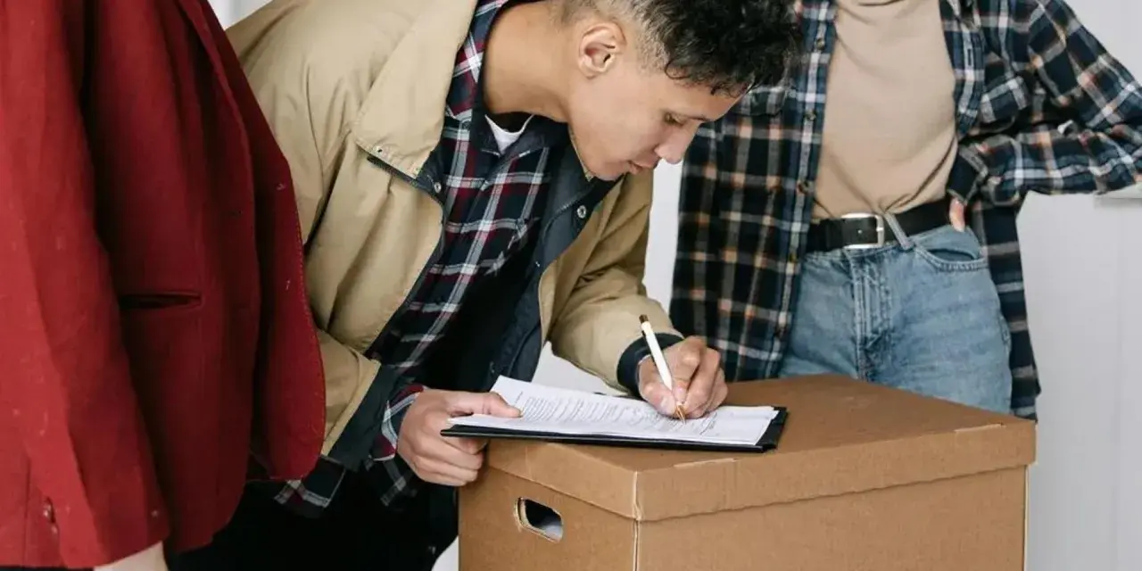 Joven firma un contrato de alquiler sobre una caja de mudanza, con otras dos personas observando.