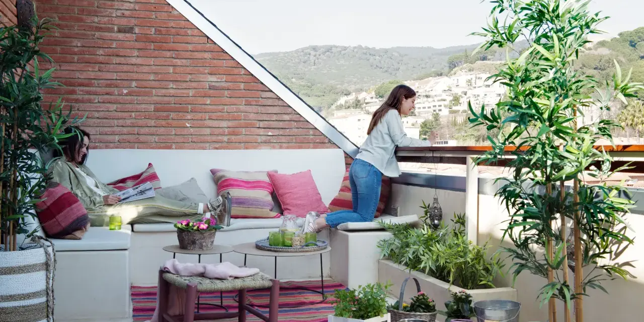 Dos mujeres disfrutan de una terraza con arboles bonitos, cojines coloridos y vistas a la montaña.