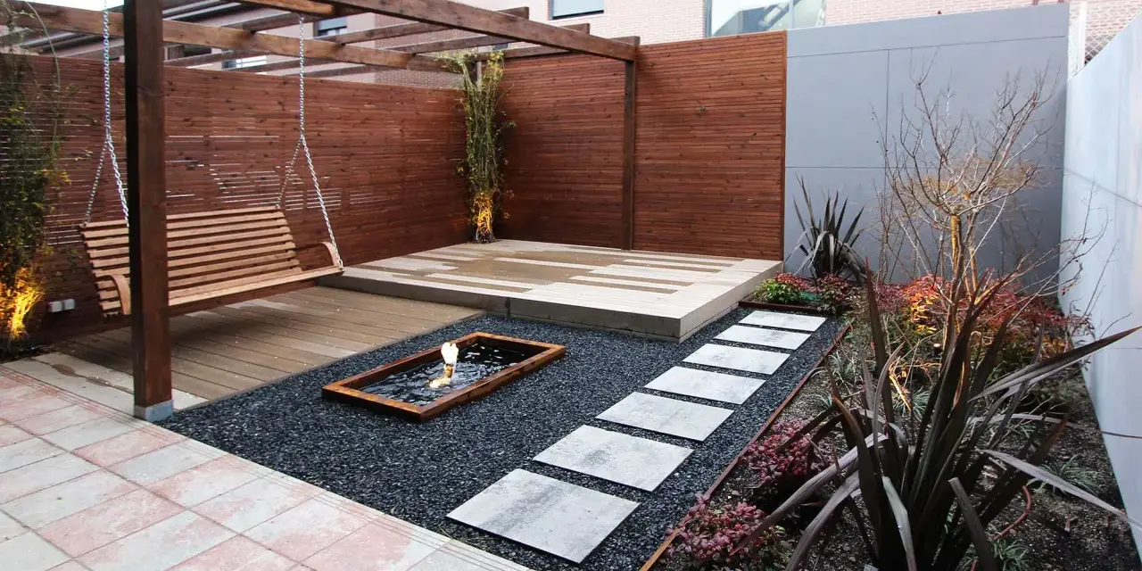 Un columpio de madera cuelga bajo un cenador. Senderos de piedra cruzan grava negra hacia un estanque. Minimalistas jardines modernos.