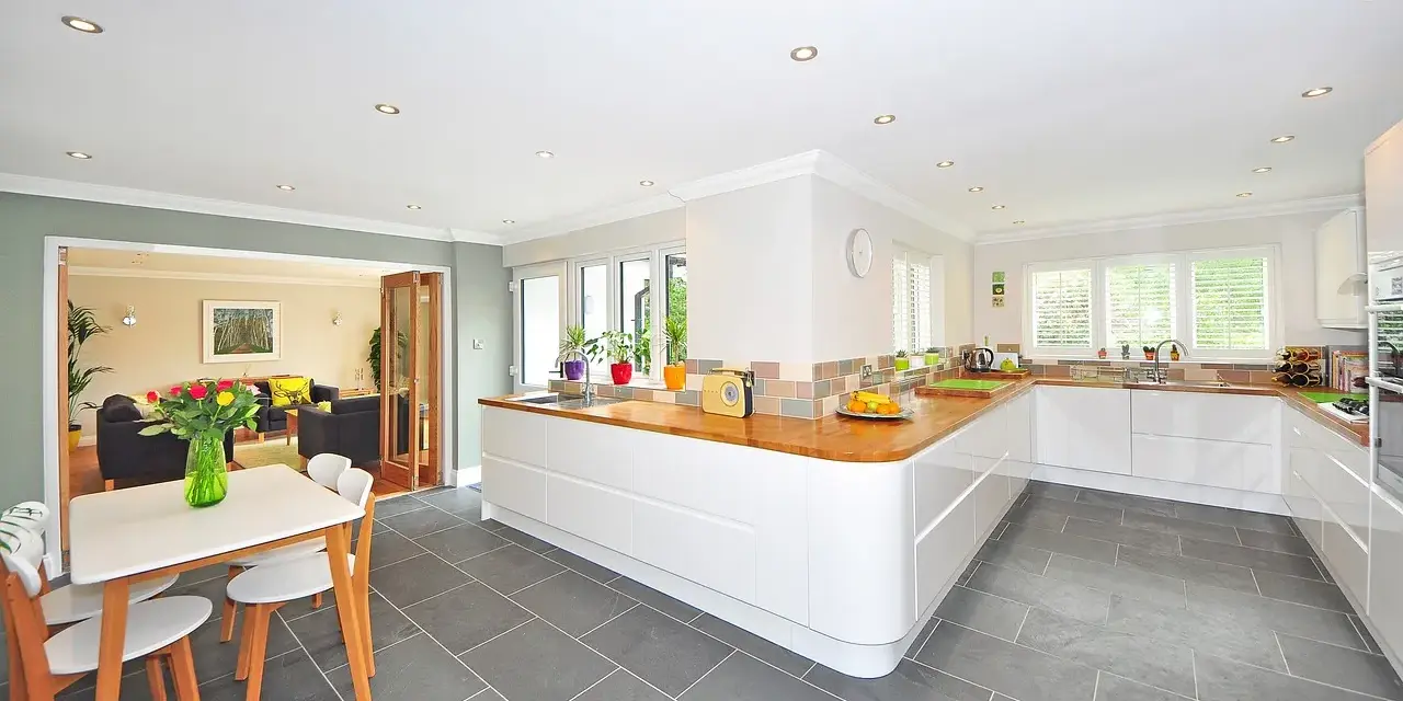 Cocina moderna con suelo gris y muebles blancos. Una mesa con sillas de madera y un jarrón con flores completan la escena.