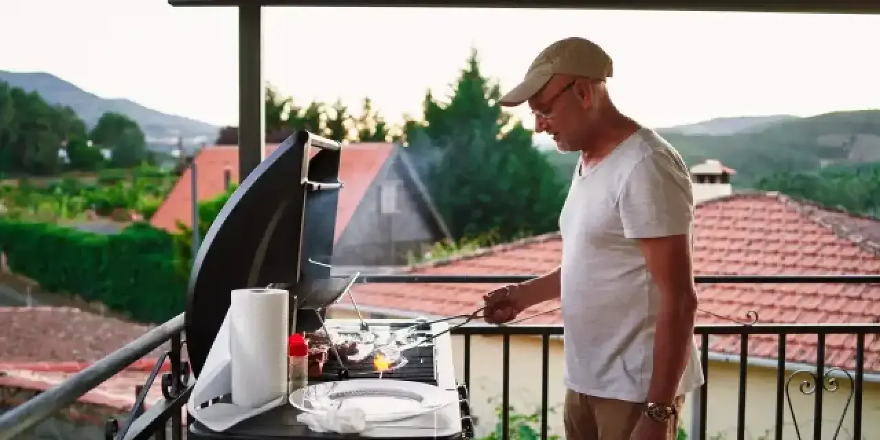 Hombre mayor cocina en barbacoa en terraza. ¡Cuidado con las multas por hacer barbacoas en terrazas!