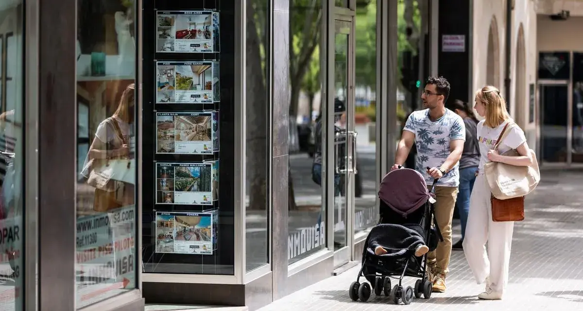 Pareja pasea con carrito, mirando escaparate inmobiliario. Quizás planean donar un piso a un hijo en Cataluña.
