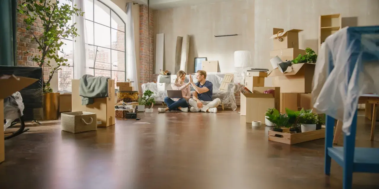 Pareja celebrando mientras planean comprar un piso a medias. Cajas y plantas por doquier, listos para empezar una nueva vida.