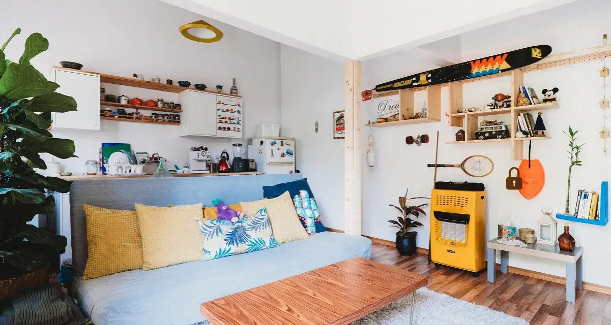 Sala de estar lista para alquilar a estudiantes. Sofá azul, mesa de centro de madera, estanterías con objetos decorativos y cocina pequeña.