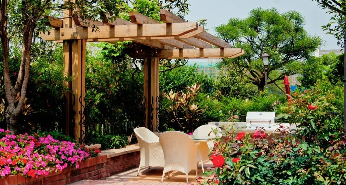 Jardín con pérgola de madera, mesa y sillas, flores rosas y rojas, y barbacoa.