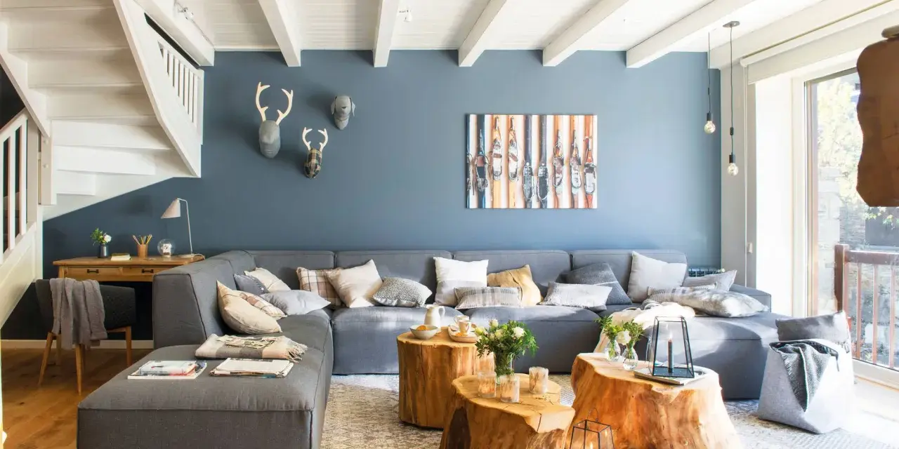 Sofá modular gris y pared azul crean un ambiente acogedor. Mesas de tronco y detalles en madera aportan calidez.
