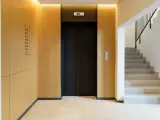 Portal moderno con ascensor negro y escaleras de mármol. Ideas para como decorar un portal de vecinos con paneles de madera y luz cálida.