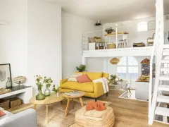 Un salón luminoso con sofá amarillo, mesas de madera y una escalera blanca que lleva a un altillo. Parece uno de esos **casas de cuento** con vistas al mar.