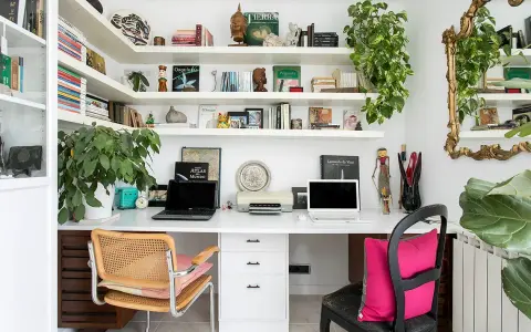 Un despacho en casa con estanterías llenas de libros y plantas. Dos portátiles, una impresora y decoración variada crean un espacio de trabajo funcional y acogedor.