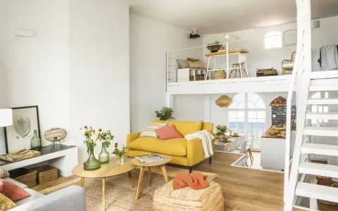 Un salón luminoso con sofá amarillo, mesas de madera y una escalera blanca que lleva a un altillo. Parece uno de esos **casas de cuento** con vistas al mar.