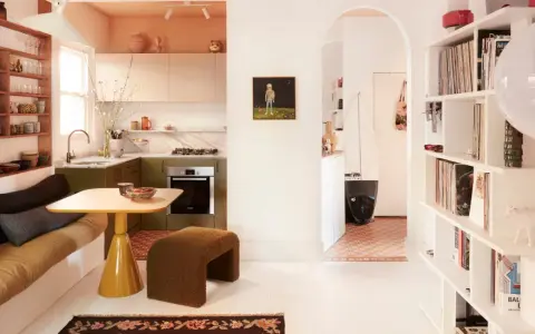 Un salón-cocina acogedor con estanterías llenas de objetos, un sofá cama y una mesa amarilla. Un pasillo arqueado conduce a otra estancia con una tabla de surf. Estas casas curiosas invitan a la vida.