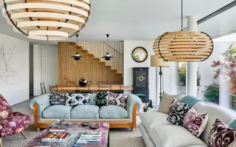 Elegante salón con sofás claros, mesa de centro con libros y lámparas de madera. El minimalismo en casa se refleja en la escalera de madera y la decoración cuidada.
