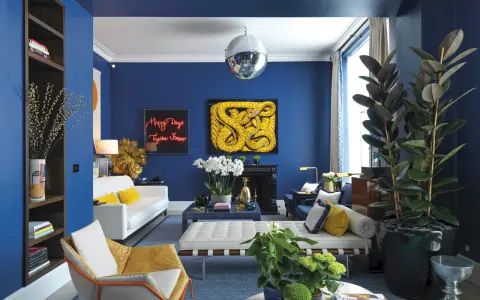 Salón moderno con paredes azul cobalto. Sofá blanco, sillón mostaza y cojines amarillos. Arte abstracto y plantas. Colores que combinan con el azul resaltan.