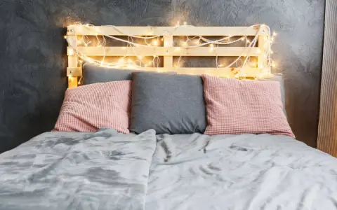 Cabecero de palets ideas: cama con cojines rosas y grises, manta gris y luces cálidas.