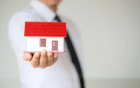 Un hombre con corbata negra sostiene una maqueta de casa blanca con techo rojo. ¿Listo para que puedas alquilar mi vivienda habitual?