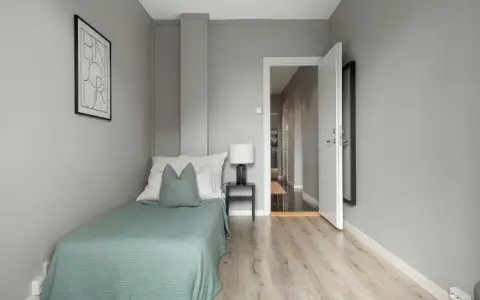 Habitación minimalista con cama individual, mesita de noche y lámpara. La puerta abierta revela un pasillo, sugiriendo que es legal alquilar habitaciones.