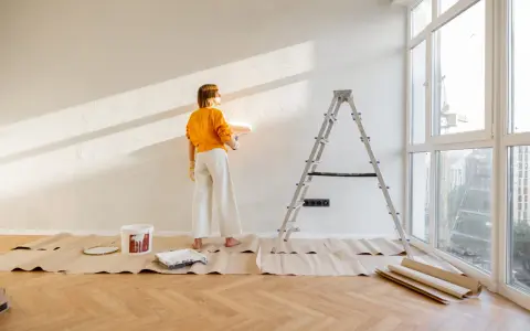 Mujer pintando pared con rodillo, lista para decorar piso antiguo de alquiler. Escalera, cubeta de pintura y protectores de suelo.
