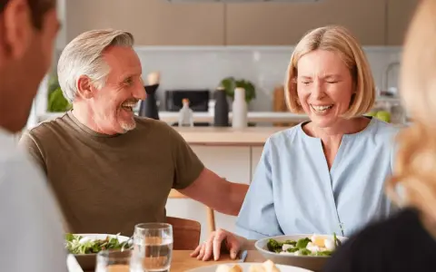 Padre e hijo ríen mientras planean comprar vivienda a medias. Comida familiar en la cocina.