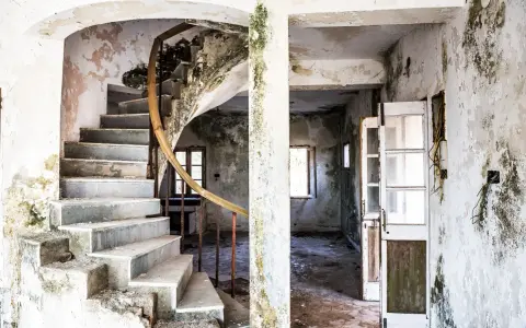 Interior de una casa en ruinas con escalera de caracol y paredes desconchadas. El estado de abandono evidencia las obligaciones del ayuntamiento para su mantenimiento.