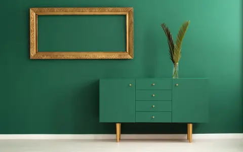Un mueble verde esmeralda con patas doradas y un marco dorado vacío sobre pared verde. Los colores que pegan con el dorado son el verde intenso y el blanco.