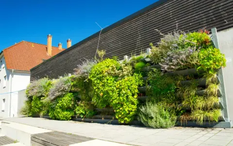 Un exuberante jardín vertical con palets y macetas, lleno de plantas verdes y flores moradas, adorna una pared de madera oscura.
