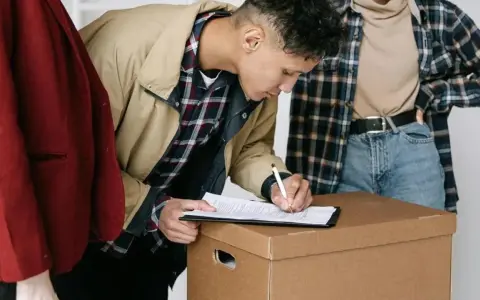 Joven firma un contrato de alquiler sobre una caja de mudanza, con otras dos personas observando.