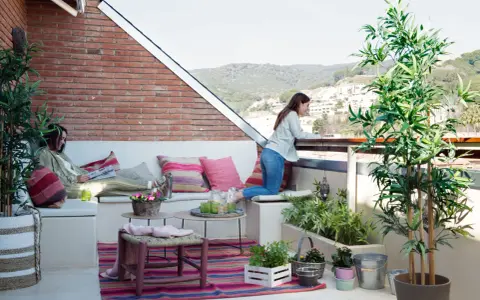 Dos mujeres disfrutan de una terraza con arboles bonitos, cojines coloridos y vistas a la montaña.