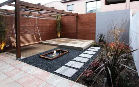 Un columpio de madera cuelga bajo un cenador. Senderos de piedra cruzan grava negra hacia un estanque. Minimalistas jardines modernos.