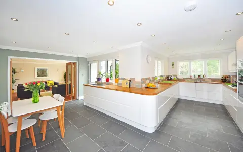 Cocina moderna con suelo gris y muebles blancos. Una mesa con sillas de madera y un jarrón con flores completan la escena.
