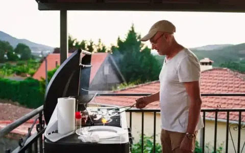 Hombre mayor cocina en barbacoa en terraza. ¡Cuidado con las multas por hacer barbacoas en terrazas!